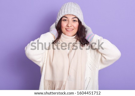 Horizontal portrait of attractive magnetic brunette putting grey hat on, wearing white sweater, scarf and gloves, looking directly at camera, standing isolated over lilac background in studio.