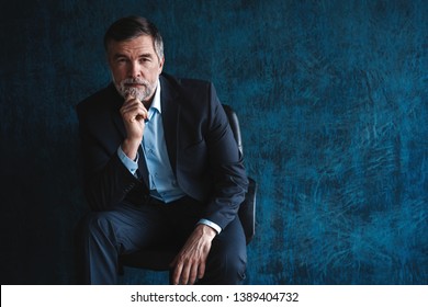 Horizontal Portrait Of Attractive Serious Mature Man Wearing Stylish Formal Clothing Sitting In Armchair And Looking At Camera Isolated Over Dark Blue Background