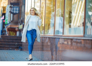 Horizontal portrait of a beautiful young caucasian blonde girl smiling and walking near shop showcase fixing her hair, blue jeans and beige trench coat, blurred blueish yellow autumn background