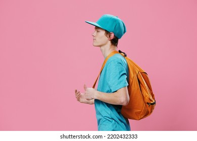 Horizontal Portrait In A Blue T-shirt And Cap With An Orange Backpack On His Back, Standing Sideways To The Camera. Studio Photo On A Plain Pink Background With Space For An Advertising Mockup