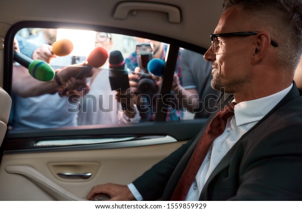 Horizontal\
portrait of calm man in suit looking at the reporters that attack\
him holding microphones in order to get exclusive interview.\
Selective focus on man. View from inside\
car