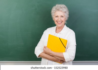 Horizontal portrait of a Caucasian cheerful female senior teacher holding a yellow file with a blank chalkboard behind - Powered by Shutterstock