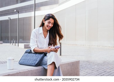 horizontal portrait of an elegant young indian woman. She is sitting near office building and using her smart phone
