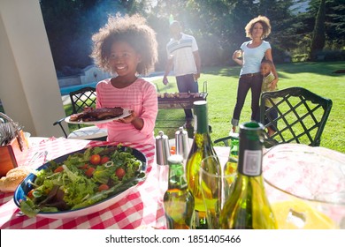 Horizontal portrait of a family of four enjoying barbeque outdoors with daughter holding a plate of meat with her family in the background.