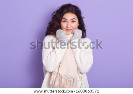 Horizontal portrait of good looking attractive young brunette putting hands on scarf, looking directly at camera, wearing white warm clothes, smiling, being in good mood. Winter holidays concept.
