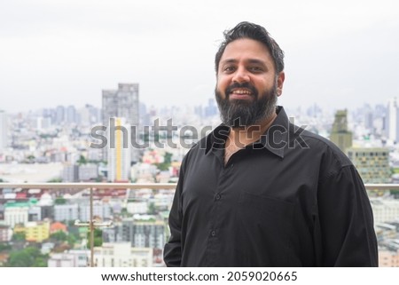 Horizontal portrait of handsome bearded Indian man smiling at rooftop in city