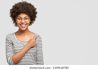Horizontal portrait of happy dark skinned female with Afro hairsyle giggles joyfully, has broad smile with white teeth, indicates aside at blank space for your advertising content. Emotions concept