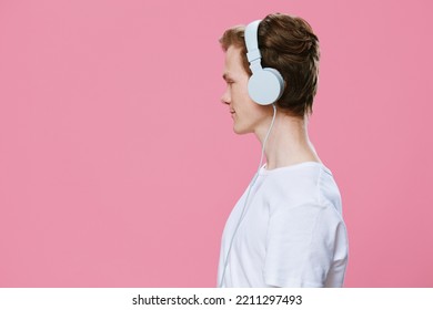 Horizontal Portrait Of A Male Student On A Pink Background Standing Sideways To The Camera In Large White Headphones And Listening To Music. Studio Photo With Empty Space For Advertising Mockup