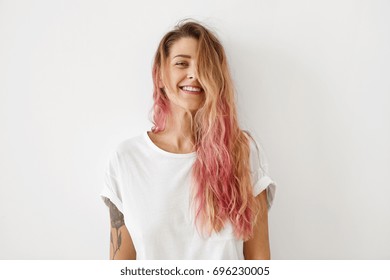 Horizontal portrait of pleasant-looking Caucasian female with long hair, pink on tips, having tattooes on arms, wearing white casual T-shirt, covering her face with hair, looking happily in camera - Shutterstock ID 696230005