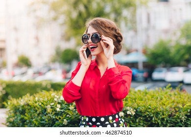 Horizontal Portrait Of Pretty Girl In Sunglasses Walking In Park. She Wears Red Blouse And Nice Hairstyle. She Is Speaking On Phone.