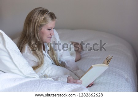 Horizontal portrait of pretty young woman with long blonde hair wearing pale casual clothes and lying on bed reading