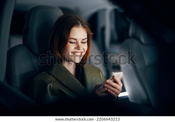 horizontal portrait of a stylish, luxurious woman in\
a green leather coat, sitting in a black car at night in the\
passenger seat, playfully biting her lower lip, holding her phone\
during the trip