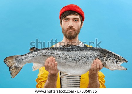 Horizontal portrait of successful angler with beard holding huge fish which he catched. Young fisherman dressed casually standing with huge trout. Man with fresh catch. Fishing and recreation