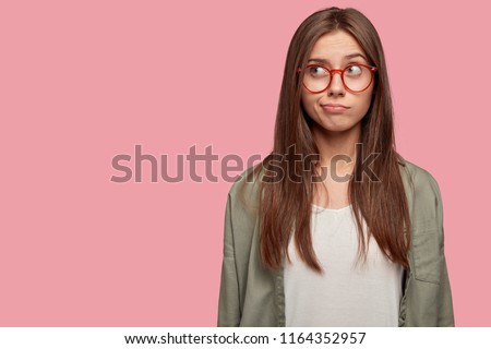 Horizontal portrait of thoughtful puzzled youngster looks with pensive dreamy expression aside, thinks what decision to make, stands against pink background with blank copy space on left side