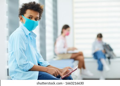 Horizontal Portrait Of Unrecognizable Black Boy Wearing Mask Sitting In Queque To School Medical Room Holding Gadget Looking At Camera, Copy Space