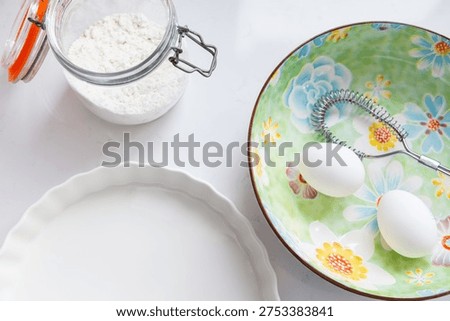 
Horizontal selective focus closeup of eggs and eggbeater in large bowl with floral motif, flour in glass jar and white ceramic pie pan on quartz counter