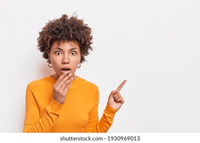 Horizontal shocked speechless woman with Afro hair keeps jaw dropped indicates away on blank space says check out something unusual wears long sleeved orange jumper isolated over white background