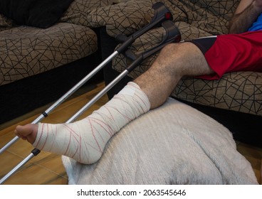 horizontal shot of an adult male with his leg in a cast and bandaged up on a foot stool with crutches lying beside him. Concept of rehabilitation of people after serious physical accident injury.