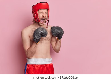 Horizontal shot of angry boxer screams loudly trains in gym prepares for sport tournament wears protective hat and gloves poses topless against pink background blank space for your promotion