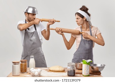 Horizontal shot of angry husband and wife feel like opponents, shoot at each other with rolling pins, cook together at home, make dough with flour, prepare delicious pastry, do bakery. Kitchen fight