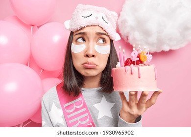 Horizontal shot of Asian woman concentrated above with upset expression applies patches under eyes feels lonely on birthday holds tasty cake dressed in soft pajama poses near inflated balloons - Powered by Shutterstock