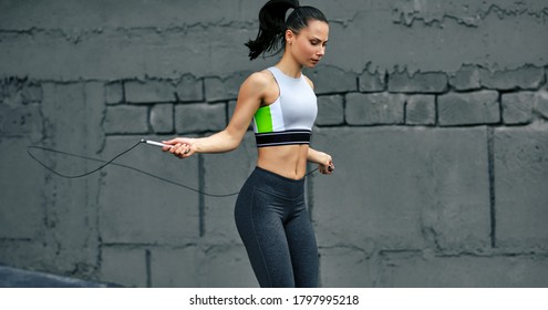 Horizontal shot of athelete determined woman skipping with rope outdoors next the cocnrete wall. Fitness female exercising with jump rope outside on a sunny day.