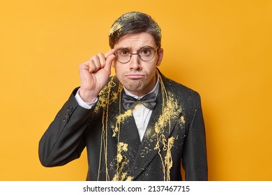 Horizontal Shot Of Attentive Serious Man Keeps Hand On Rim Of Spectacles Dressed In Festive Suit Smeared With Serpantine Spray Has Bad Mood Isolated Over Yellow Background Upset To Get Older