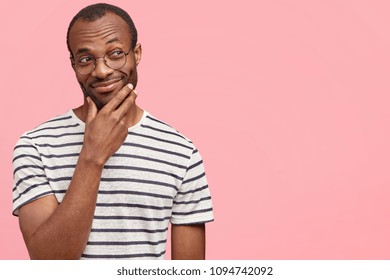 Horizontal shot of attractive dark skinned male with satisfied expression holds hand above chin, looks thoughtfully and happily aside, tries to focus or think up plan, stands over pink wall.
