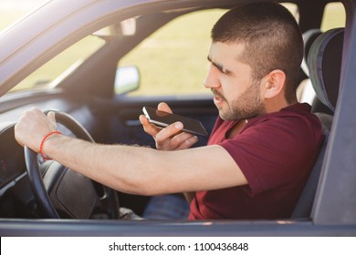 Horizontal shot of attractive male driver being in car, stuck in traffic jam, holds smart phone, speaks with girlfriend during driving, enjoys communication, motion, speed and modern technology