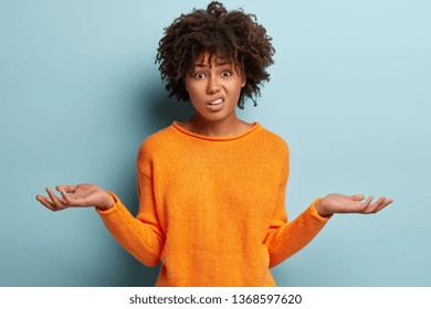 Horizontal shot of attractive woman with puzzled expression, spreads hands with confused look, frowns face, wears orange clothes, feels hesitant, indignant isolated over blue background. Doubt concept - Shutterstock ID 1368597620