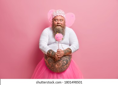 Horizontal Shot Of Bearded Fatso Man Foolishes Around On Childrens Birthday Party, Holds Magic Wand Wears Fairy Costume, Has Fat Tattooed Stomach, Isolated Over Pink Background. Happy Festival