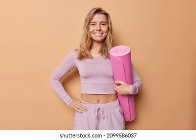Horizontal shot of beautiful blonde woma has slim fit body dressed in top and shorts holds rolled fitness mat leads healthy lifestyle poses indoor against beige background prepares for yoga practice