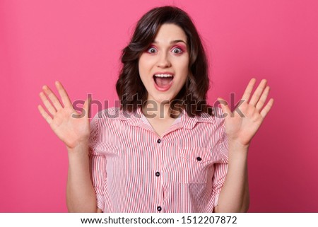 Horizontal shot of beautiful caucasian woman with shocked facial expression, keeps hands up, having widely opened eyes and mouth, has dark hair, wearing casual shirt, posing against pink wall.