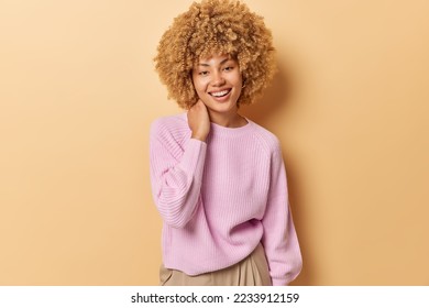 Horizontal shot of beautiful curly haired young woman smiles toothily keeps hand on neck wears casual pink jumper looks delighted at camera isolated over beige background. Happy emotions concept