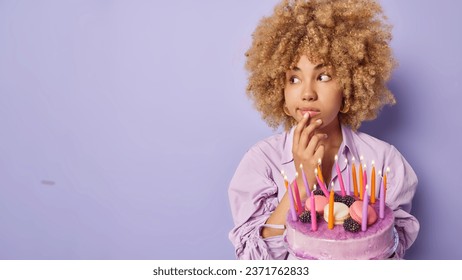 Horizontal shot of beautiful curly haired woman keeps hand on chin concentrated aside poses with tasty cake and burning candles celebrates special occasion isolated over purple background copy space