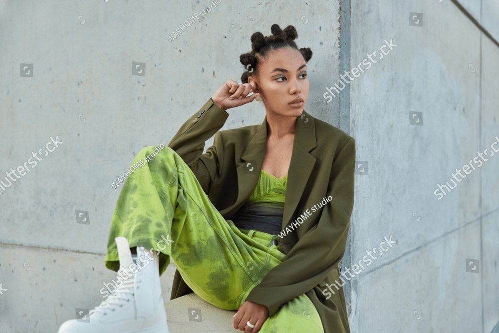 Horizontal shot of beautiful teenage girl with trendy hairstyle dressed in green clothes looks away poses against urban grey wall considers something. People youth lifestyle and fashion concept