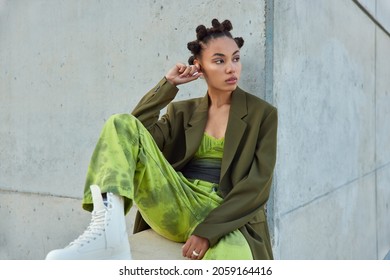 Horizontal shot of beautiful teenage girl with trendy hairstyle dressed in green clothes looks away poses against urban grey wall considers something. People youth lifestyle and fashion concept - Shutterstock ID 2059164416