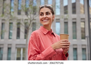 Horizontal Shot Of Beautiful Young Woman Smiles Happily Enjoys Leisure Time Walks Outside With Disposabe Cup Of Coffee Wears Pink Shirt Glasses On Head Being In Good Mood Strolls Against Urban Setting