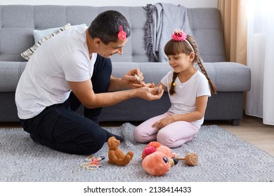 Horizontal Shot Of Brunette Funny Man Wearing White T Shirt Sitting On Floor And Playing With His Daughter, Father Painting Child Nails, Spending Time Together.