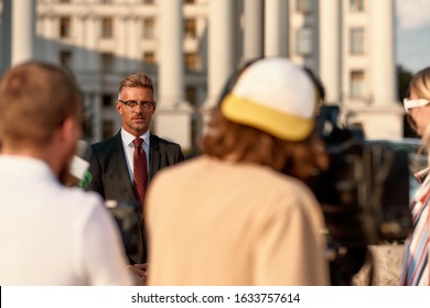 Horizontal Shot Of Businessman Giving Interview To Journalists. People Making Interview Using Digital Camera And Equipment Set At Outdoor Location. Selective Focus