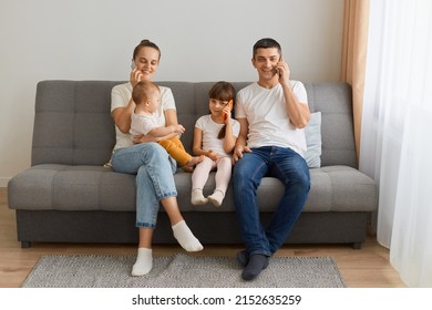 Horizontal Shot Of Busy Family Sitting On Sofa And Having Conversation On Smart Phone, Looking At Camera, Kid With Smart Phone Talking Too, Family Spending Time With Gadgets At Home.