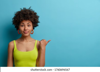 Horizontal shot of calm curly haired Afro American woman points thumb away, wears casual green vest, shows space for your advertisement, isolated on blue background. People, advertisement concept - Shutterstock ID 1760696567