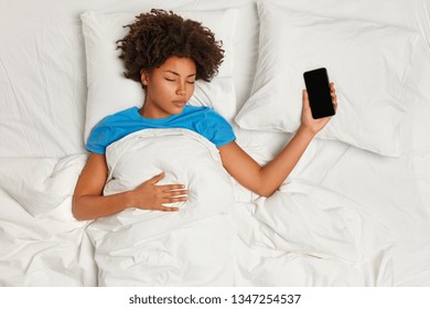 Horizontal Shot Of Calm Restful Dark Skinned Woman Falls Asleep With Modern Smart Phone, Sleeps Well, Lies Under White Duvet, Waits For Call From Boyfriend. Sleeping, Technology, People, Rest Concept