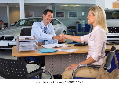 Horizontal shot of a car salesman and female customer sitting at the desk and shaking hands in a large car showroom. - Powered by Shutterstock