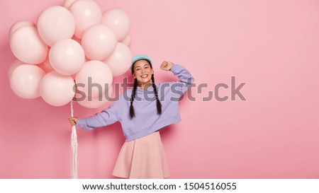 Horizontal shot of carefree happy Asian girl celebrates first day at college, carries big bunch of pink inflated balloons, wears blue cap, oversized jumper and skirt, poses over rosy wall, empty space