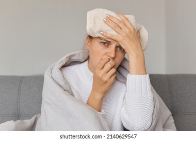 Horizontal Shot Of Caucasian Ill Woman Being Wrapped In Blanket Sitting On Sofa With Wet Towel On Her Forehead, Looking At Camera With Upset Face, Suffering Headache, Runny Nose And High Temperature.