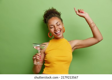 Horizontal Shot Of Cheerful Curly Haired Woman Dances Carefree Wears Yellow T Shirt And Earrings Has Pink Manicure Holds Glass Of Cocktail Isolated Over Vivid Green Background Has Fun On Party.