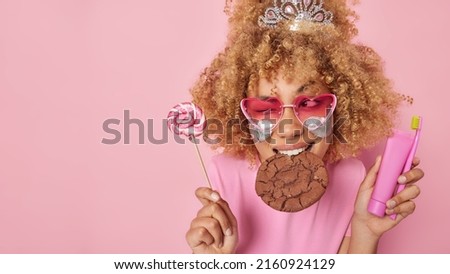 Horizontal shot of cheerful curly haired woman eats tasty cookie winks and looks gladfully aside holds lollipop in one hand and teeth care product in other isolated over pink background empty space