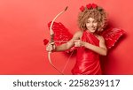 Horizontal shot of cheerful female cupid shoots arrow targets on something wears red dress with wings behind back looks mysteriously somewhere isolated over red background copy space for your text