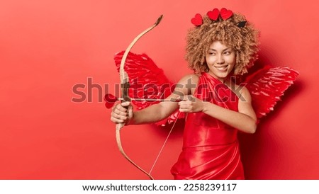 Horizontal shot of cheerful female cupid shoots arrow targets on something wears red dress with wings behind back looks mysteriously somewhere isolated over red background copy space for your text